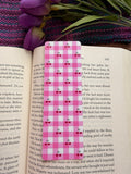 Pink checkered bookmark on an open book with a purple flower and green leaves.